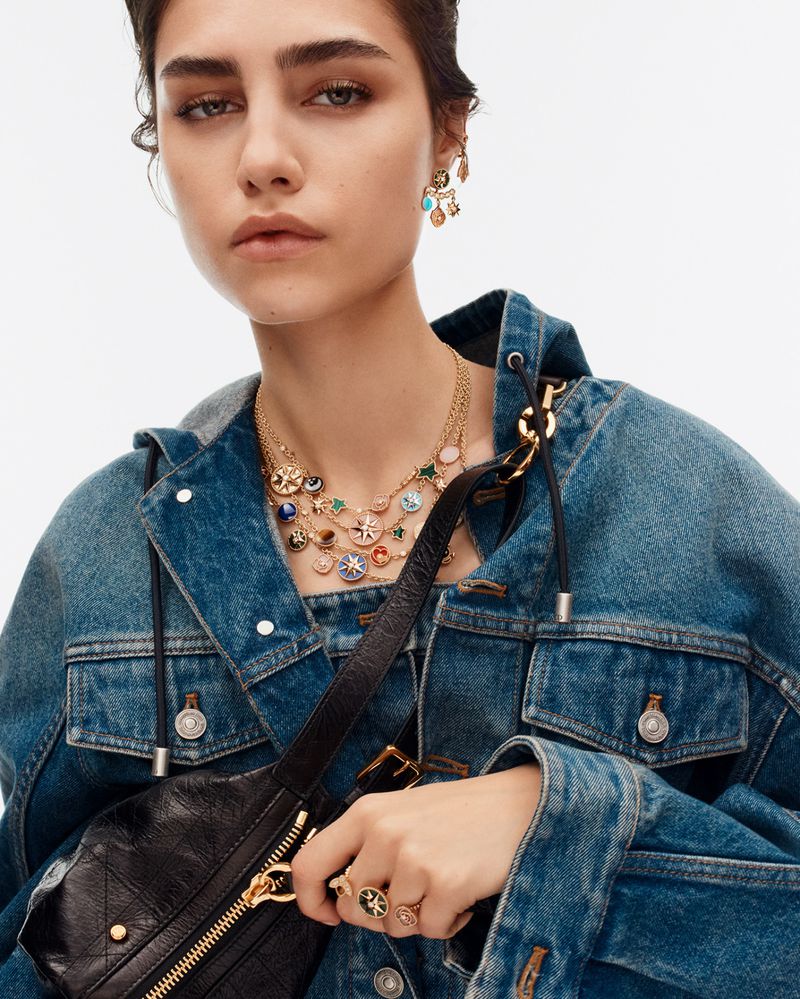 Close-up of a woman in a denim jacket wearing earrings, a necklace, and a ring, posing in a brightly lit, neutral indoor setting.