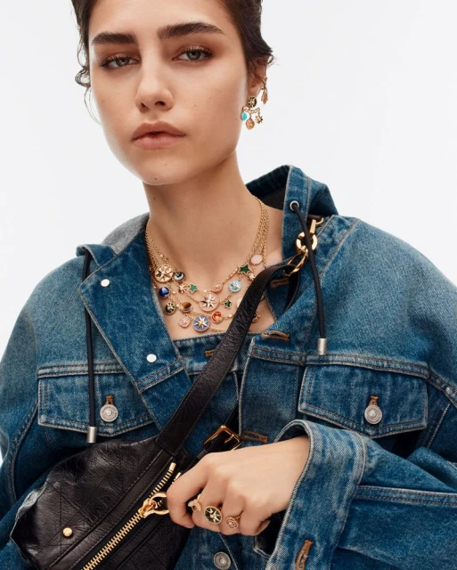 Close-up of a woman in a denim jacket wearing earrings, a necklace, and a ring, posing in a brightly lit, neutral indoor setting.