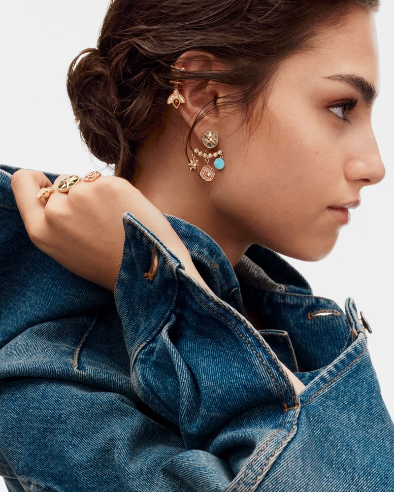 Model wearing gold earrings and rings in a close-up fashion shot, wearing a blue denim jacket against a white backdrop.