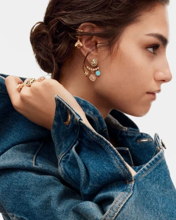 Model wearing gold earrings and rings in a close-up fashion shot, wearing a blue denim jacket against a white backdrop.