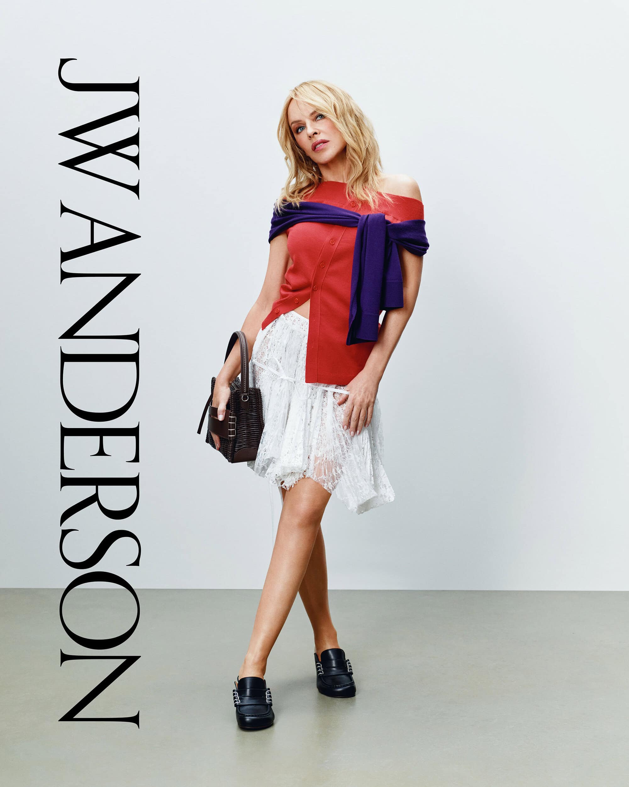 Kylie Minogue wearing an off-the-shoulder red top and white skirt holding a dark handbag against a grey studio floor and white wall