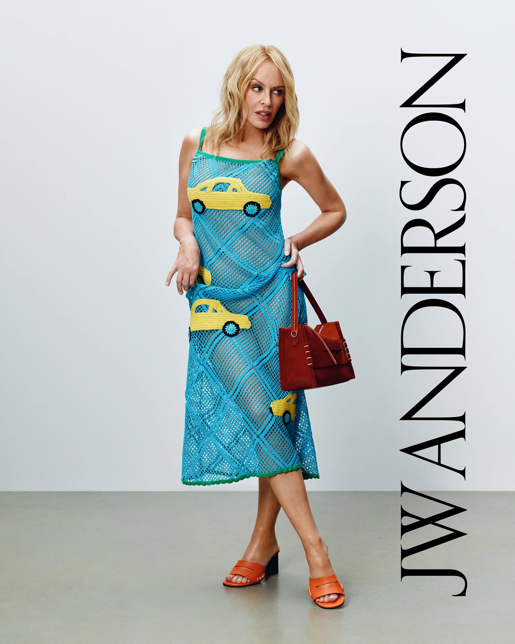 Kylie Minogue wearing a blue knit dress with yellow car motifs holding a red leather handbag against a plain background