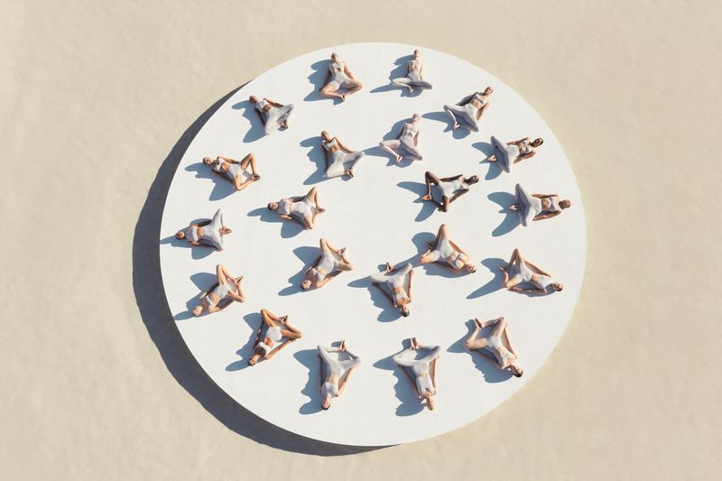 Overhead shot showcases a circular arrangement of miniature human figures in various yoga poses on a white surface against a cream-colored backdrop.
