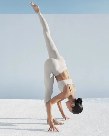 Woman in neutral activewear performing a standing split pose outdoors against a minimal wall and clear blue sky.