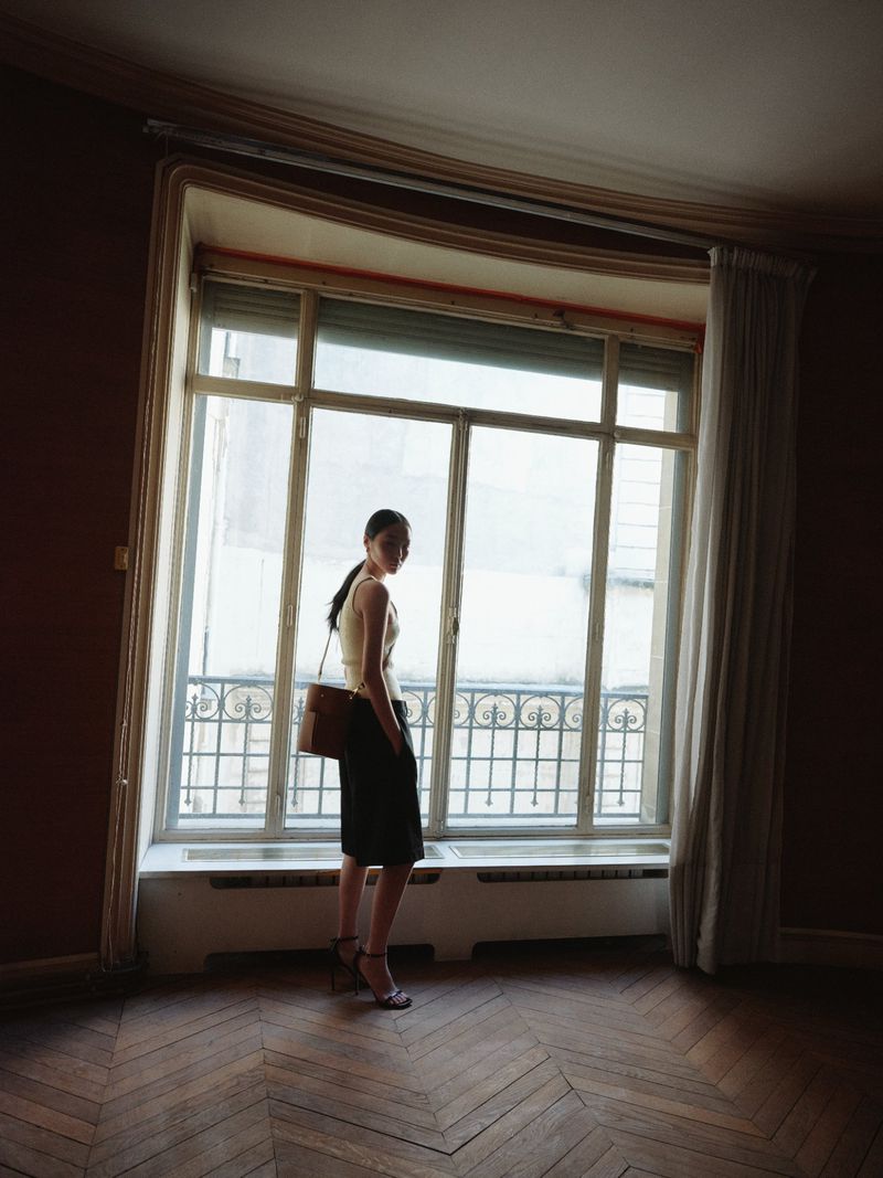 Elegant woman stands in empty room, looking out large window. She wears a skirt, top, heels, and a shoulder bag in natural light.
