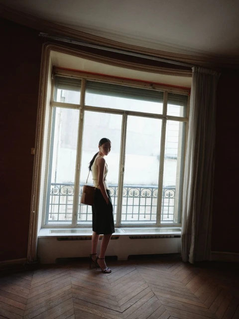 Elegant woman stands in empty room, looking out large window. She wears a skirt, top, heels, and a shoulder bag in natural light.