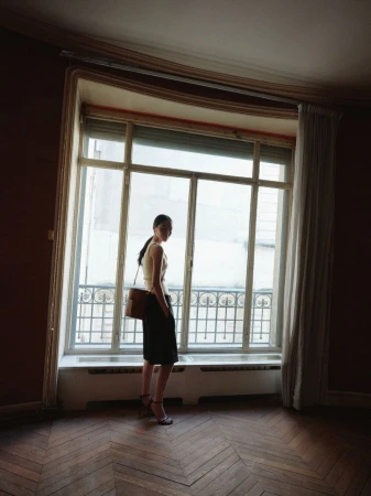 Elegant woman stands in empty room, looking out large window. She wears a skirt, top, heels, and a shoulder bag in natural light.