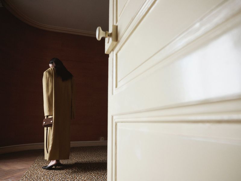 Back view of a woman in a long coat holding a handbag, standing on a patterned floor near a door, in an indoor setting.