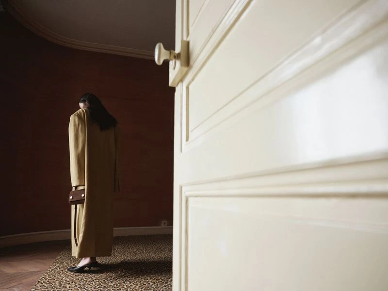 Back view of a woman in a long coat holding a handbag, standing on a patterned floor near a door, in an indoor setting.