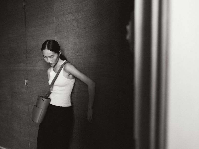 Monochrome portrait of an Asian woman in a white tank top, with a crossbody bag, standing against a textured wall in soft lighting.