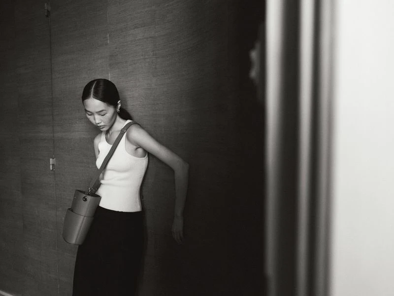 Monochrome portrait of an Asian woman in a white tank top, with a crossbody bag, standing against a textured wall in soft lighting.