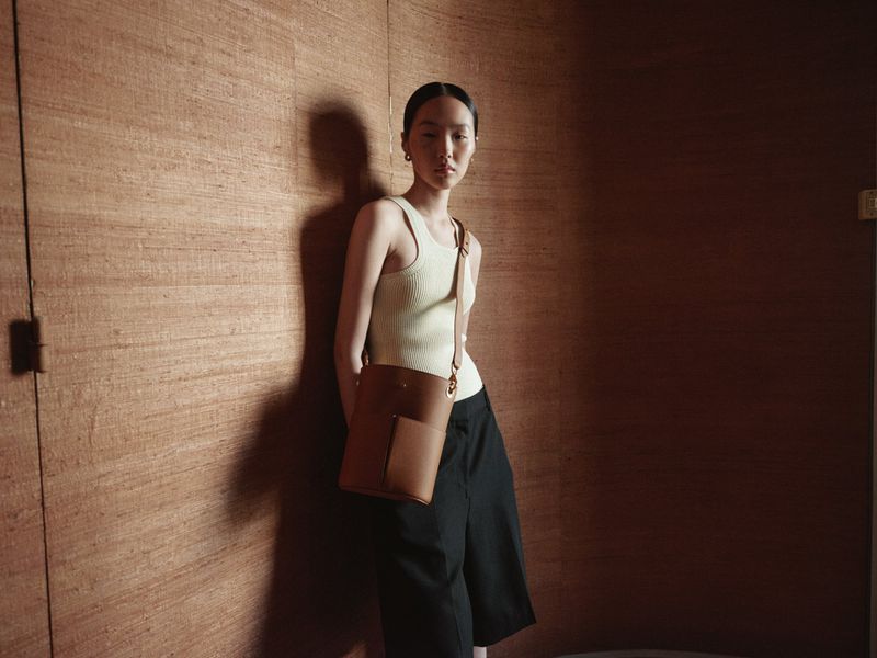 A stylish young woman with a tan leather bucket bag stands against a textured brown wall in a fashion or lifestyle setting.