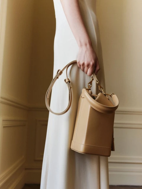 Cream colored purse with gold hardware on a woman's shoulder with a white dress against a neutral colored wall in a studio.