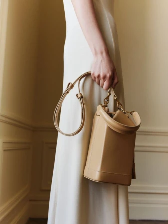 Cream colored purse with gold hardware on a woman's shoulder with a white dress against a neutral colored wall in a studio.