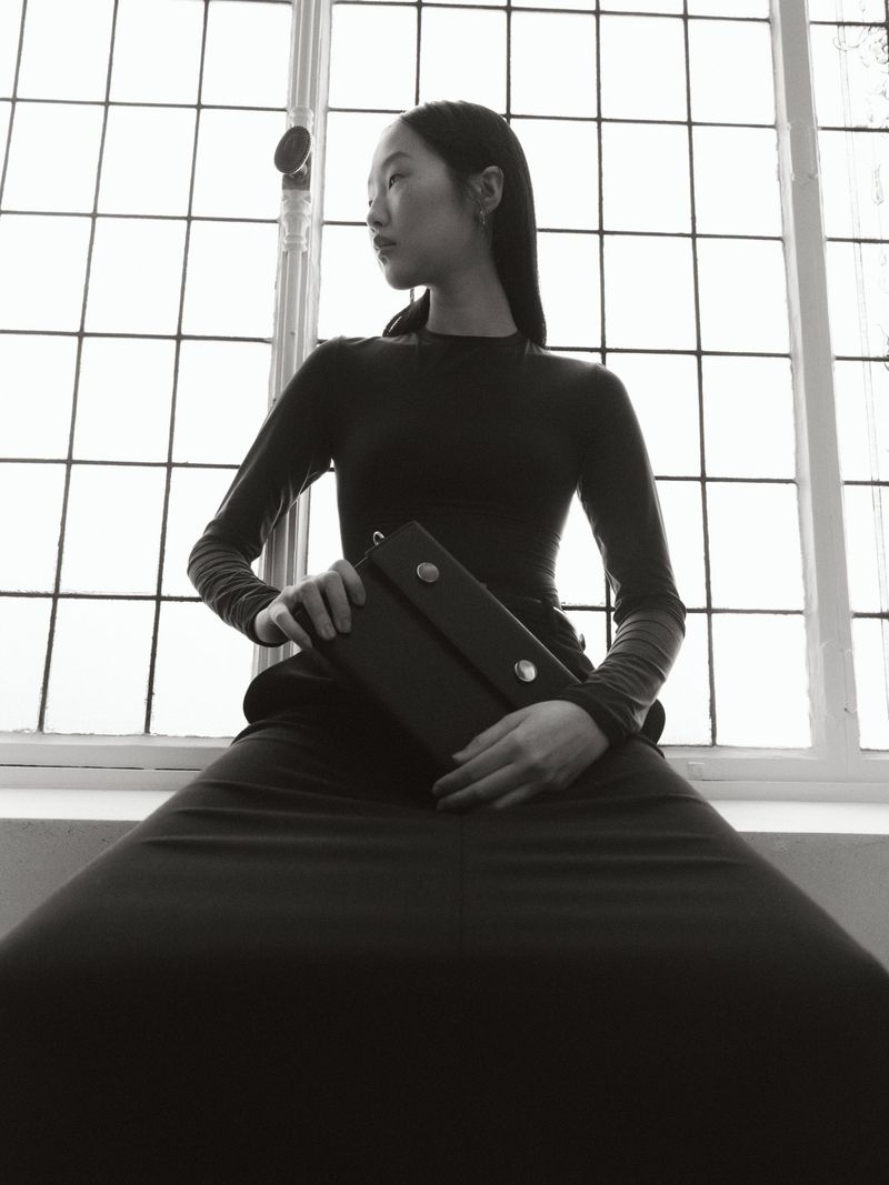 Black-and-white fashion portrait of an Asian woman seated in front of a window, holding a dark, rectangular clutch. Classic, editorial.