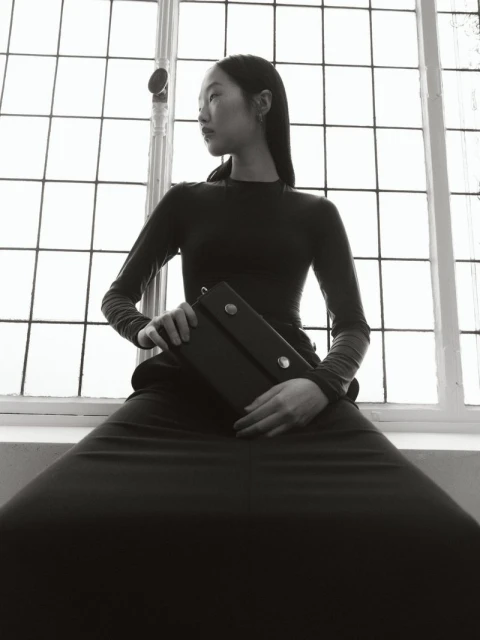 Black-and-white fashion portrait of an Asian woman seated in front of a window, holding a dark, rectangular clutch. Classic, editorial.