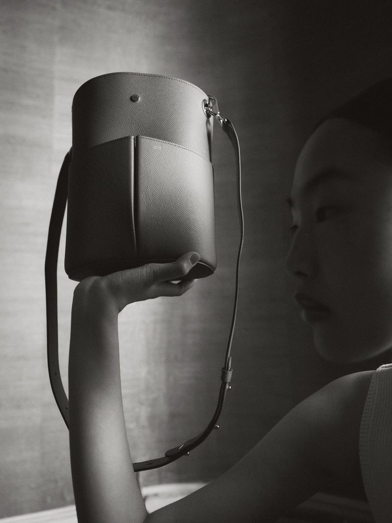 A woman in a neutral setting holds a stylish cylindrical bucket bag with a shoulder strap. The image is in black and white.