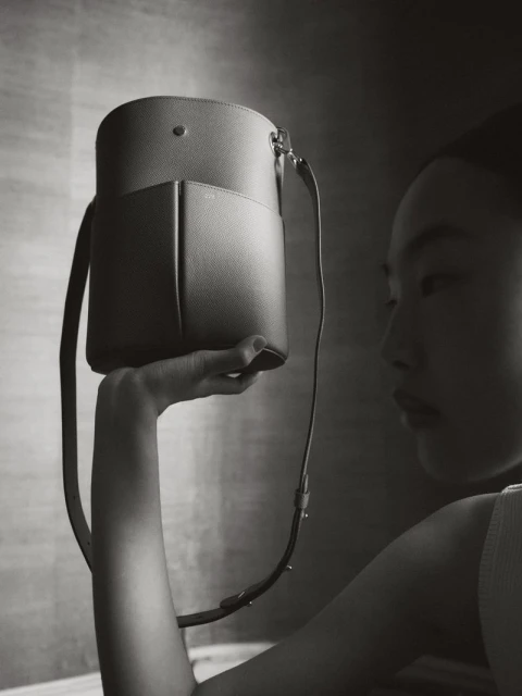 A woman in a neutral setting holds a stylish cylindrical bucket bag with a shoulder strap. The image is in black and white.