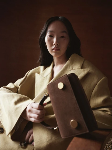 Portrait of an Asian woman in a khaki jacket holding a brown suede handbag with gold hardware against a warm brown background.