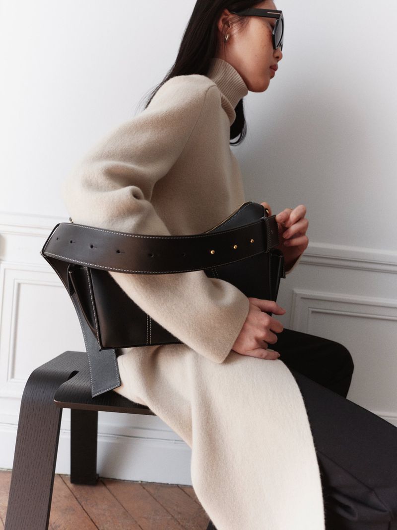 Elegant woman in a cream-colored sweater dress sits on a modern black chair, holding a black leather bag against a white wall.