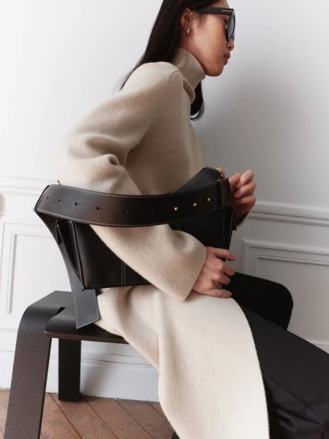 Elegant woman in a cream-colored sweater dress sits on a modern black chair, holding a black leather bag against a white wall.