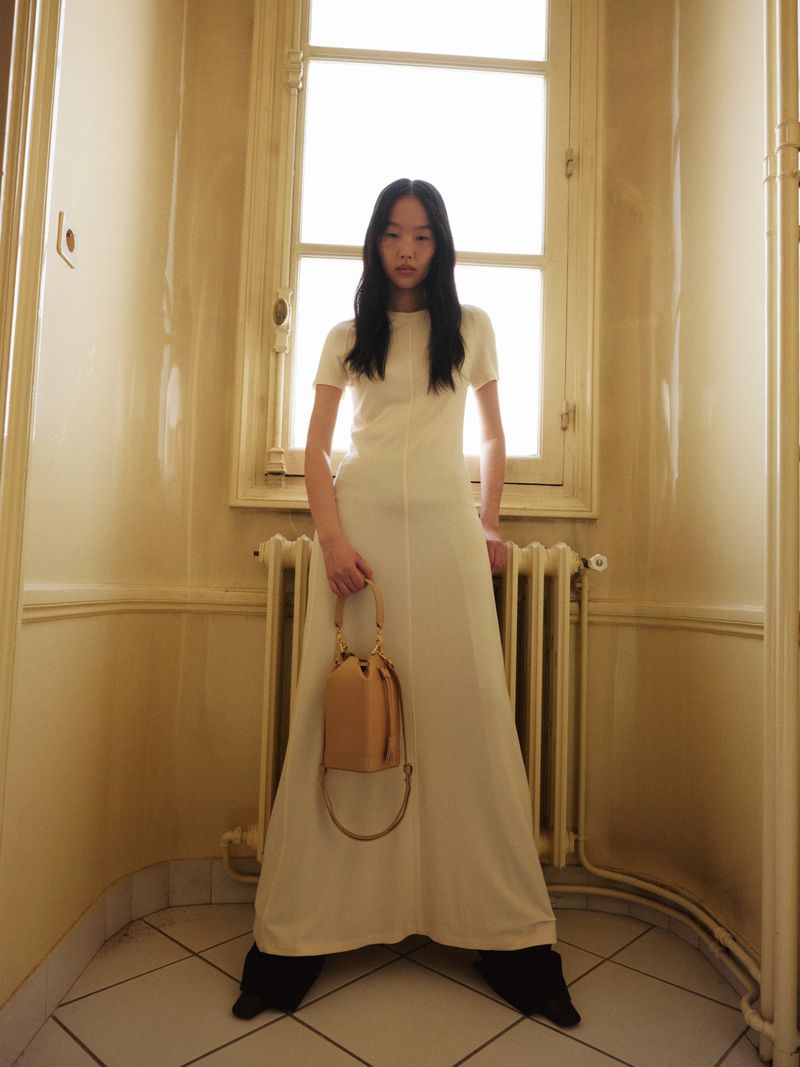 A woman in a white dress stands in a hallway holding a handbag, bathed in soft, diffused light from a window behind her.