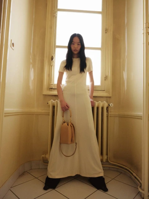 A woman in a white dress stands in a hallway holding a handbag, bathed in soft, diffused light from a window behind her.