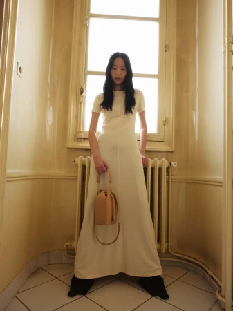 A woman in a white dress stands in a hallway holding a handbag, bathed in soft, diffused light from a window behind her.