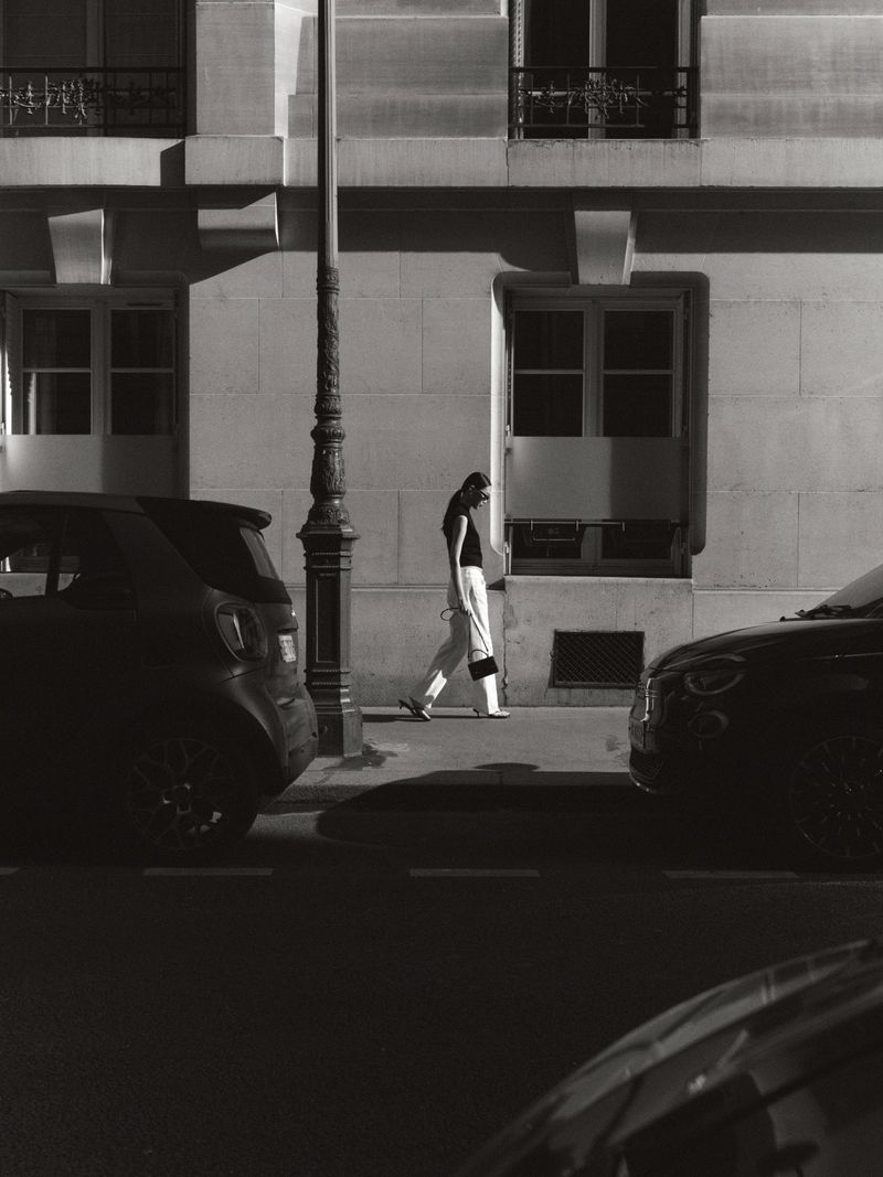Monochrome image of a woman walking along a sidewalk next to parked cars and a building with windows and a street light.