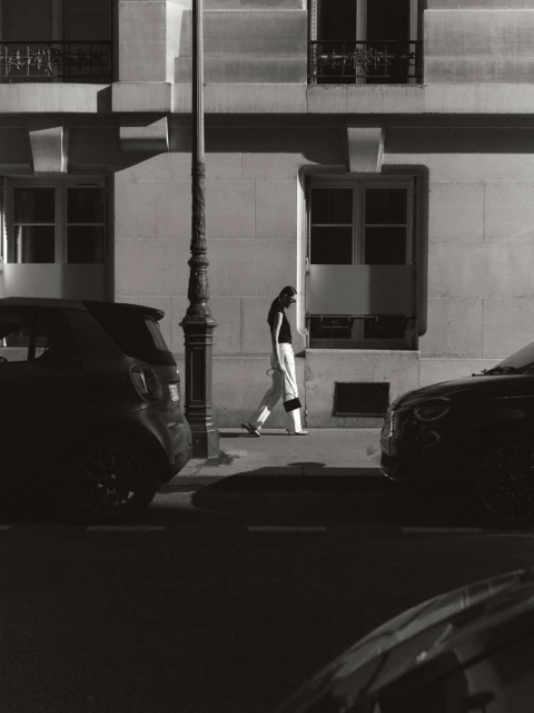 Monochrome image of a woman walking along a sidewalk next to parked cars and a building with windows and a street light.