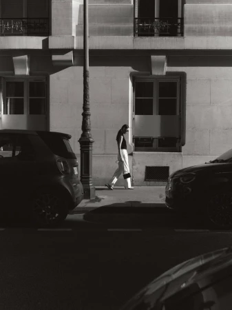 Monochrome image of a woman walking along a sidewalk next to parked cars and a building with windows and a street light.