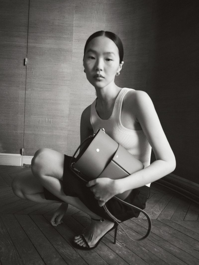 Elegant Asian woman squats indoors wearing a tank top and shorts, holding a satchel-style purse, accessorized with heels and hoop earrings.