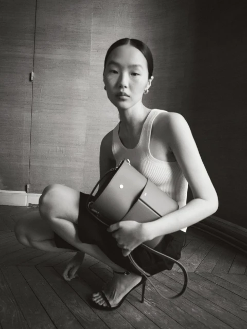 Elegant Asian woman squats indoors wearing a tank top and shorts, holding a satchel-style purse, accessorized with heels and hoop earrings.