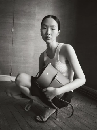 Elegant Asian woman squats indoors wearing a tank top and shorts, holding a satchel-style purse, accessorized with heels and hoop earrings.