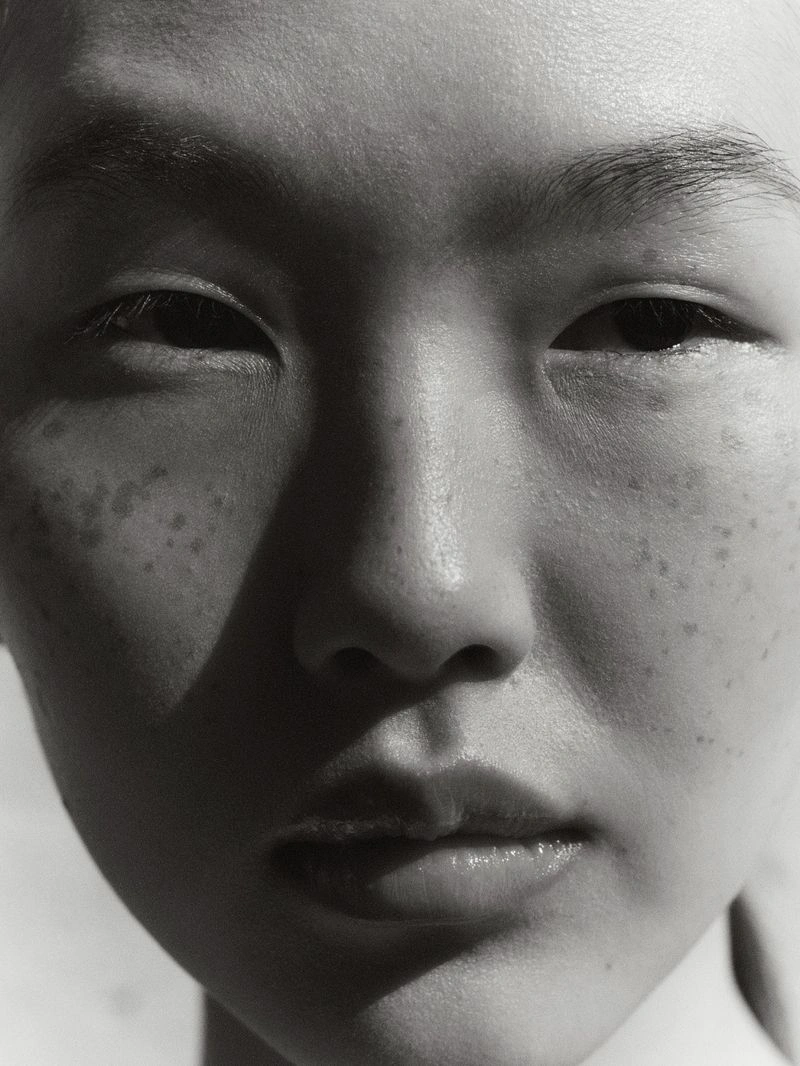 Close-up monochrome portrait of a model featuring natural skin texture, freckles, and strong dramatic directional lighting.