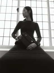 Model Peng Chang holding a leather handbag against a grid window for RSVP Paris, photographed in monochrome.