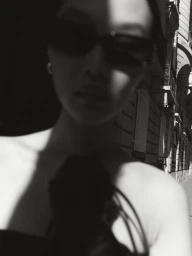 Black and white portrait of a model wearing dark sunglasses outdoors on a city street with strong shadows.