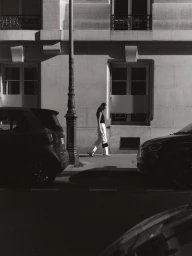 Monochrome street-level fashion editorial image showing a model walking past parked cars in front of a classical building facade.