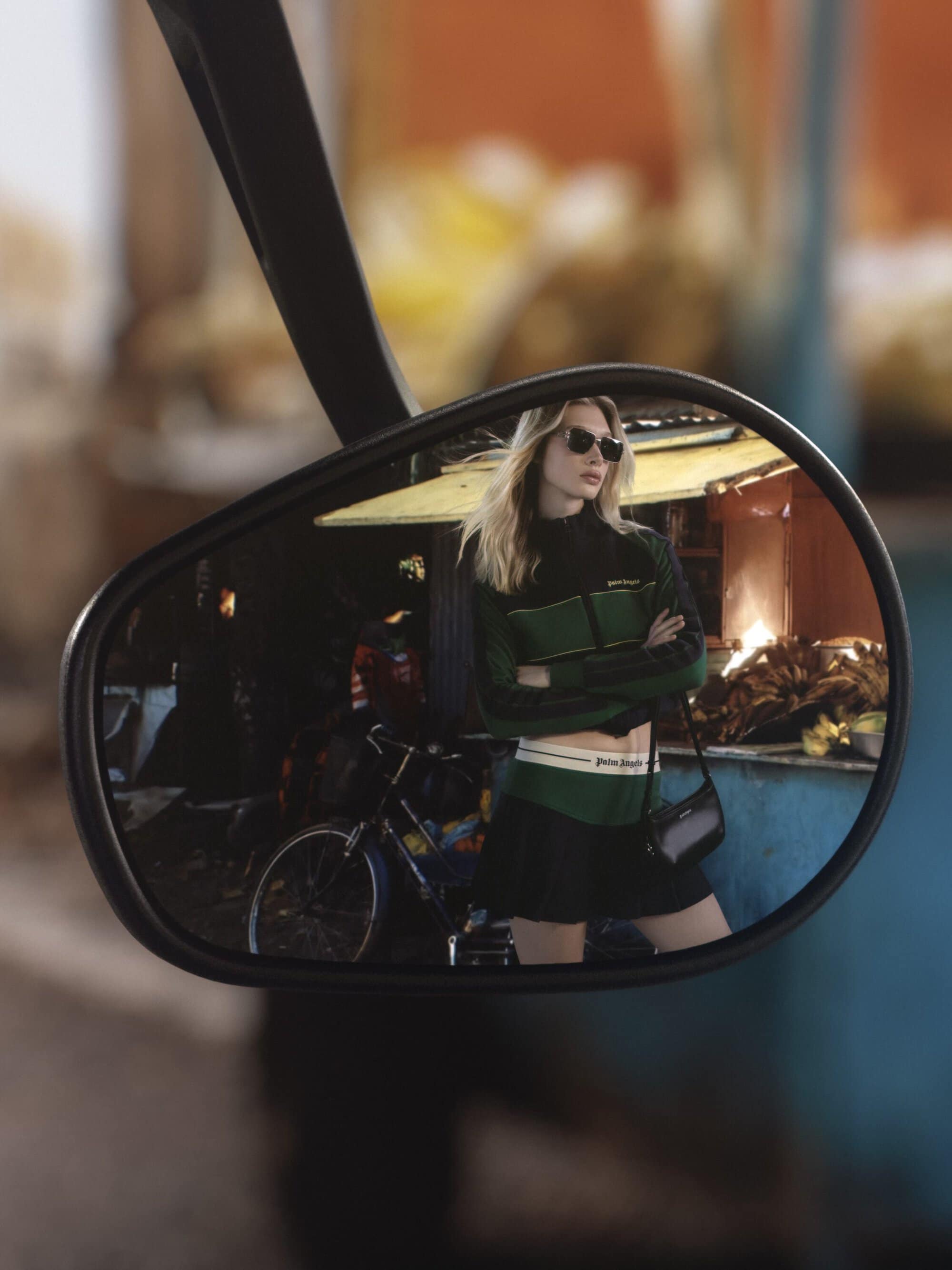 A woman wearing sunglasses and a green athletic top reflected in a motorcycle rearview mirror outdoors.