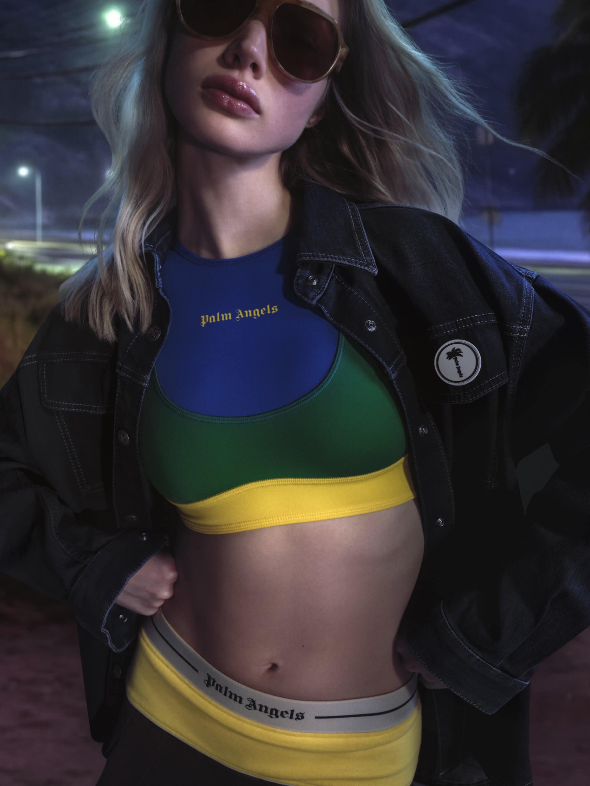 Model wearing dark sunglasses, multi-colored sports crop top, and dark denim jacket outdoors at night.