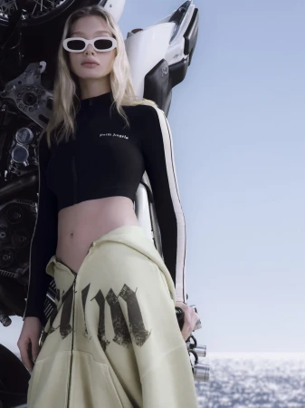 Model in white sunglasses and black long-sleeve crop top standing in front of a motorcycle against a clear sky background