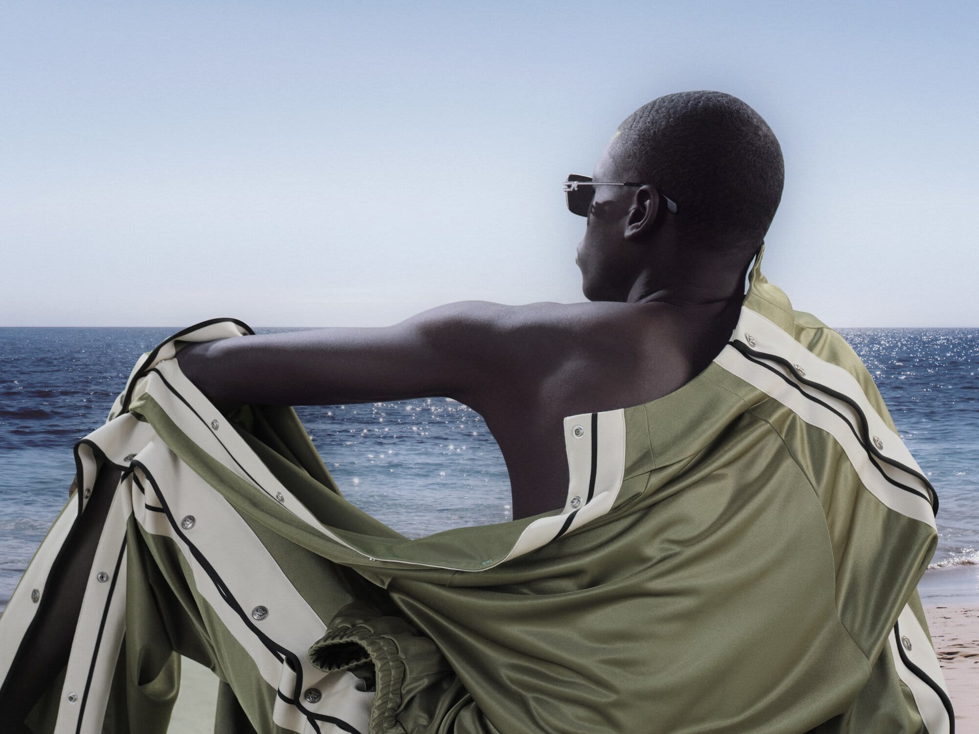 Man wearing sunglasses and olive green track jacket sitting by the ocean for Palm Angels Island Speed campaign