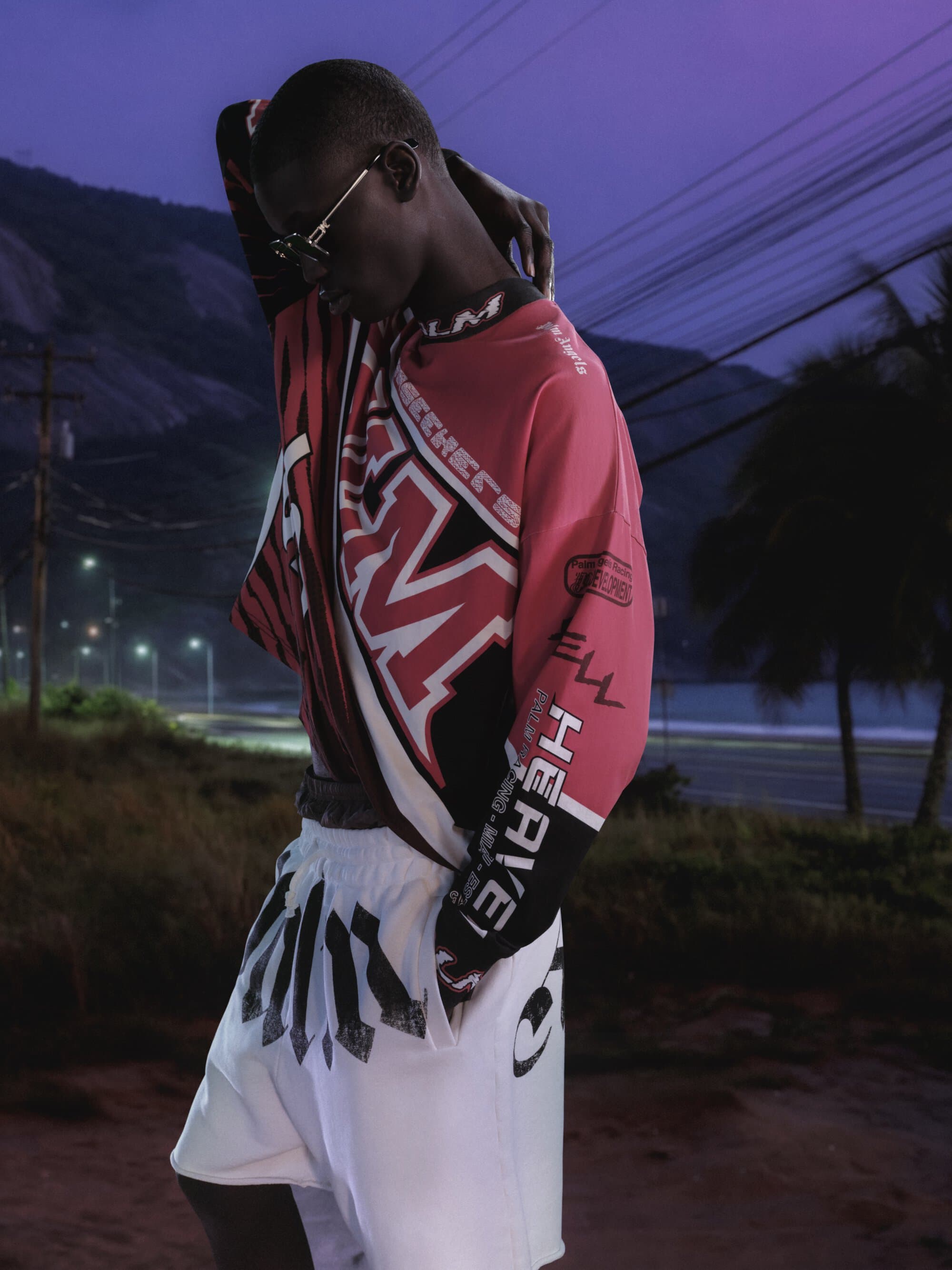 Black man wearing printed long-sleeve jersey and white shorts outdoors at dusk under a purple sky