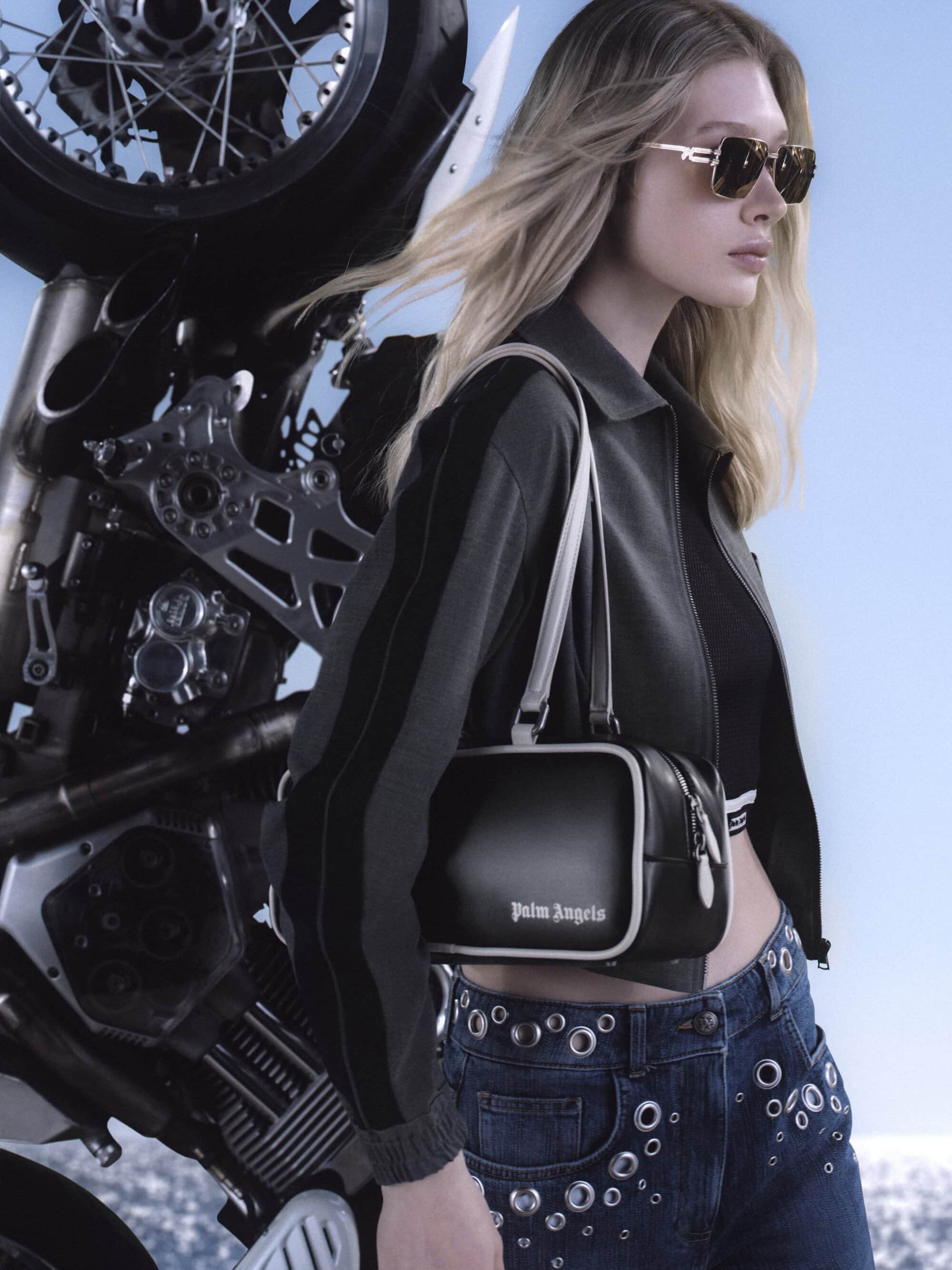 Blonde female model wearing sunglasses and black jacket standing beside a motorcycle against a sky background