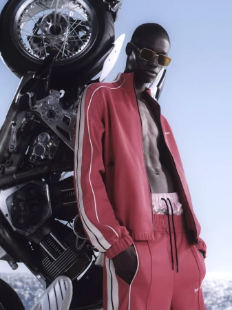 Black man wearing a red tracksuit and sunglasses leaning against a motorcycle with a clear sky in the background