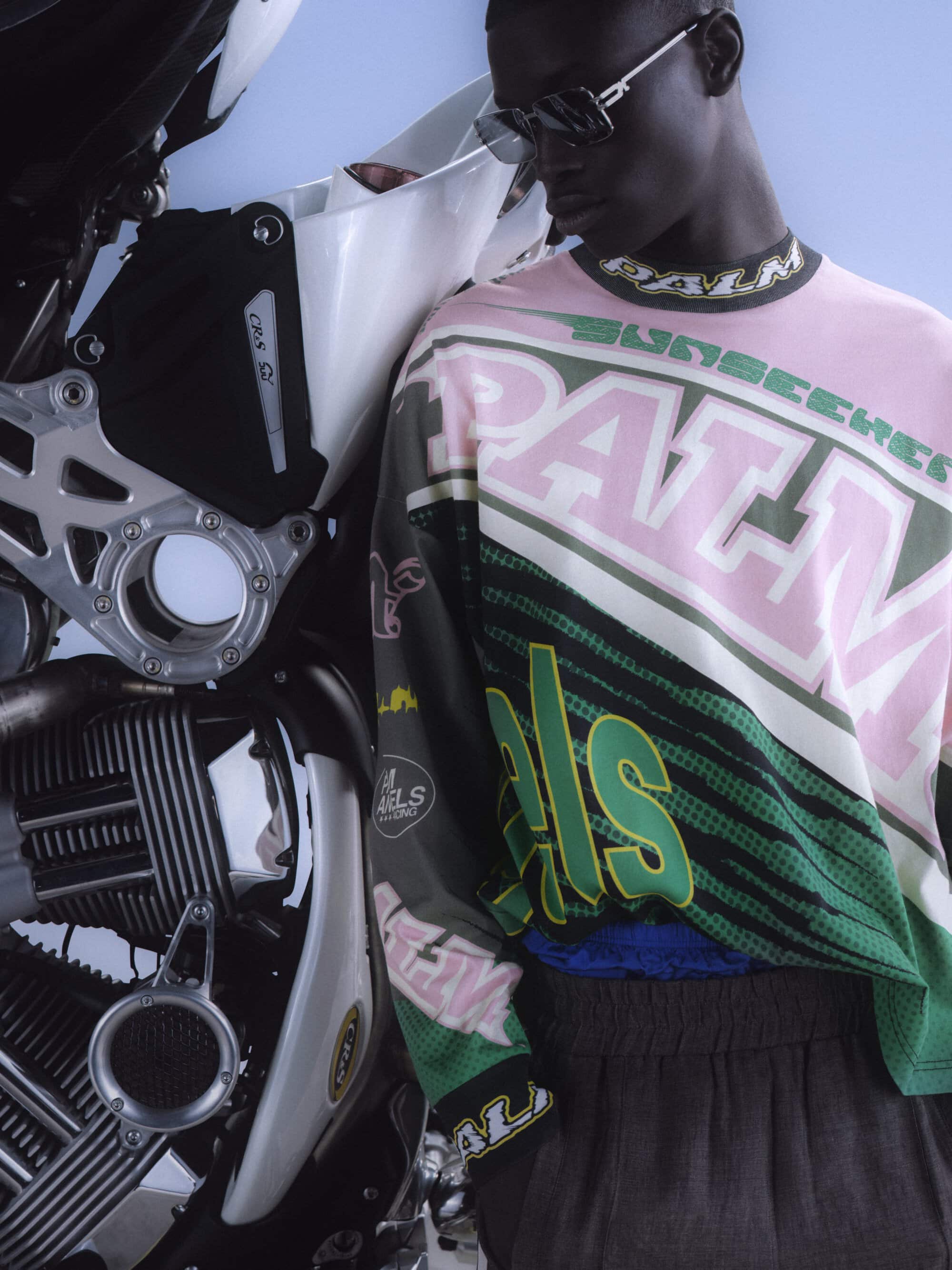 Black man wearing sunglasses and a patterned long-sleeve top leaning against a motorcycle with clear sky background