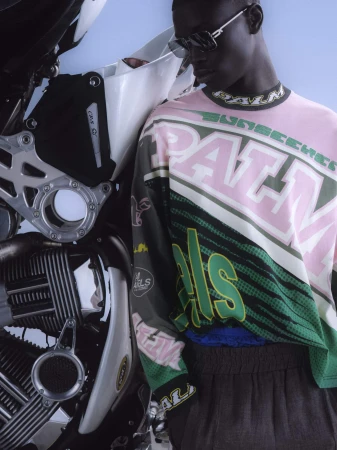 Black man wearing sunglasses and a patterned long-sleeve top leaning against a motorcycle with clear sky background