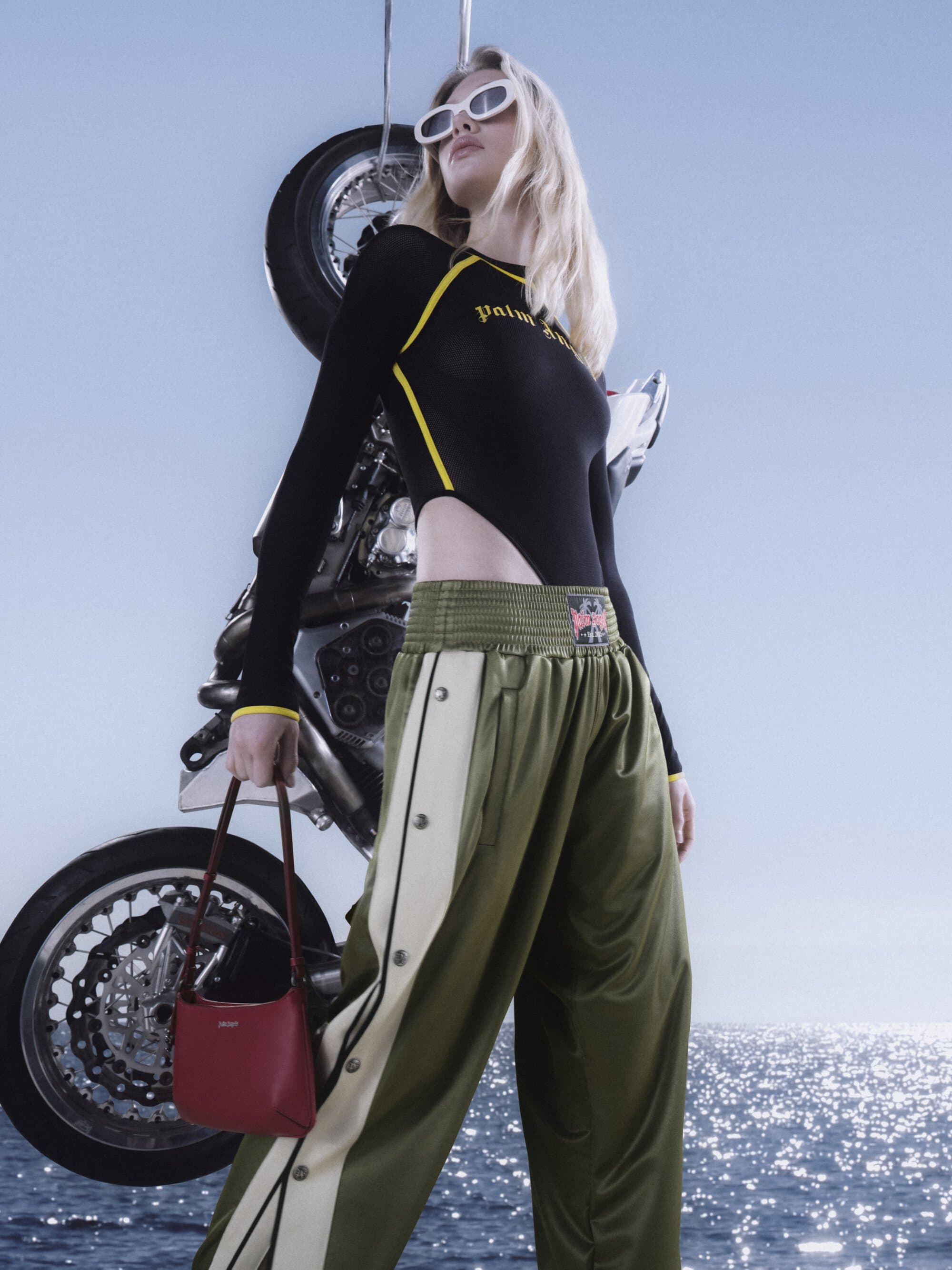 Model in white sunglasses wearing a black bodysuit and green track pants holding a small red handbag against a motorcycle and sea