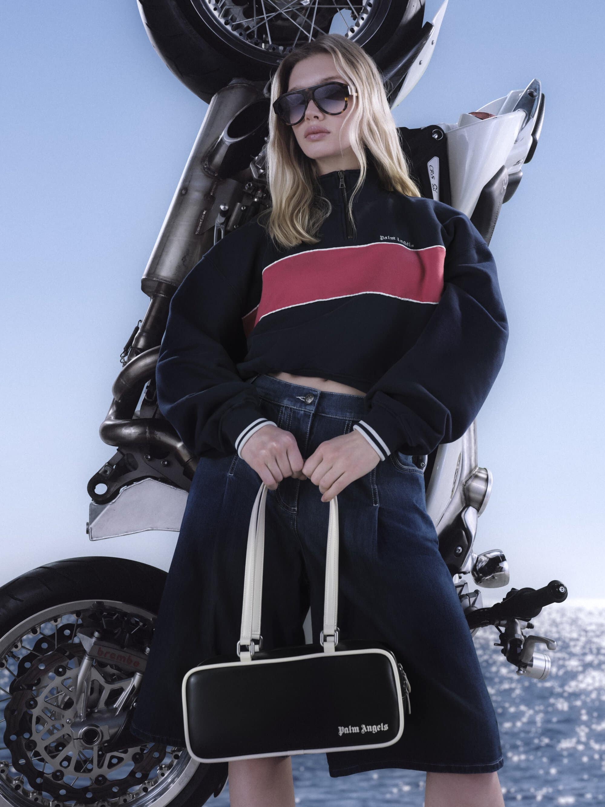 Model in sunglasses wearing a dark sweatshirt and denim skirt holding a black handbag against a motorcycle and sea.