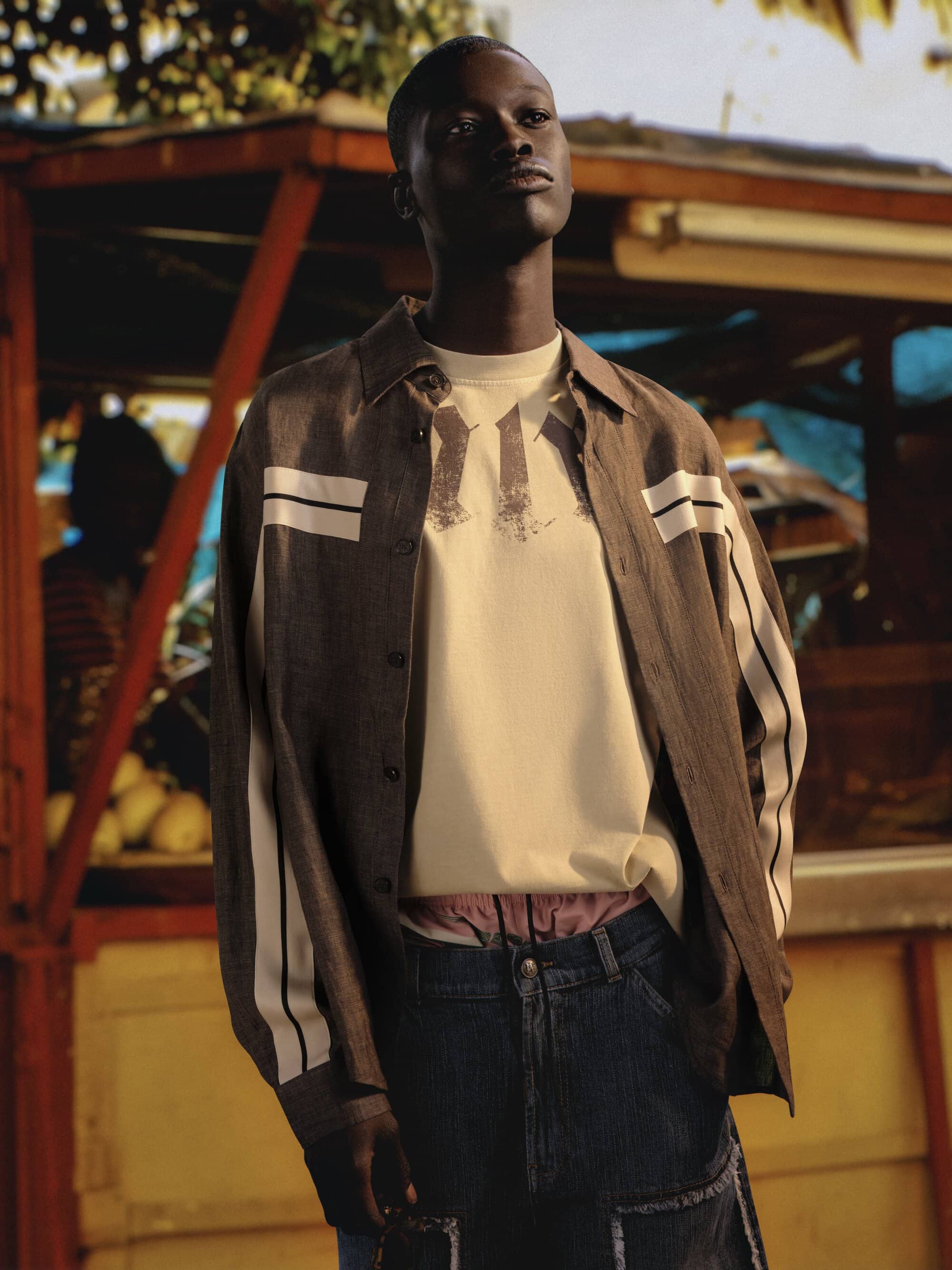 Black male model wearing a striped button-up shirt over a cream t-shirt and denim jeans outdoors.